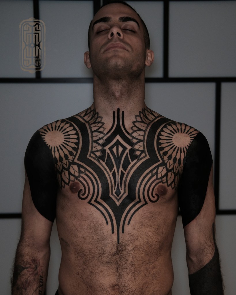 A man with a full chest tattoo featuring intricate geometric and floral designs, standing with his eyes closed against a minimalist background.