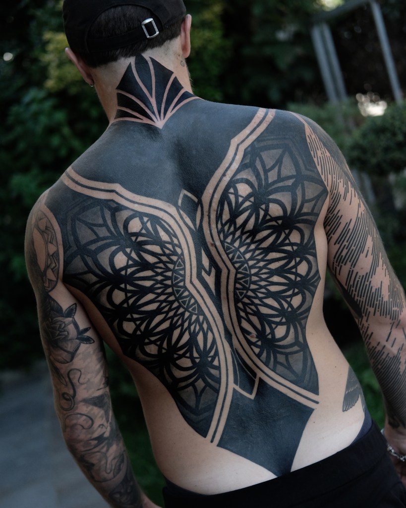 A person showing a large, intricate black tattoo on their back, featuring geometric patterns and a cover up tattoo.