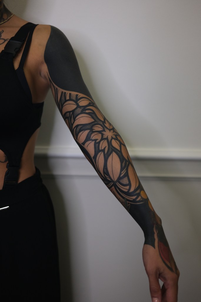 Close-up of a person's arm featuring intricate tattoo designs, with dark ink and highlights of skin tone visible. A big bold chrysanthemum flower is the main subject-