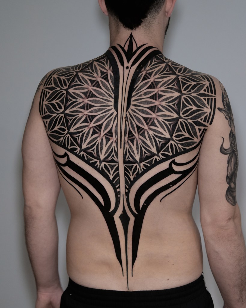 A close-up of a large and intricate black tattoo design covering the back of a person, featuring geometric and floral patterns.