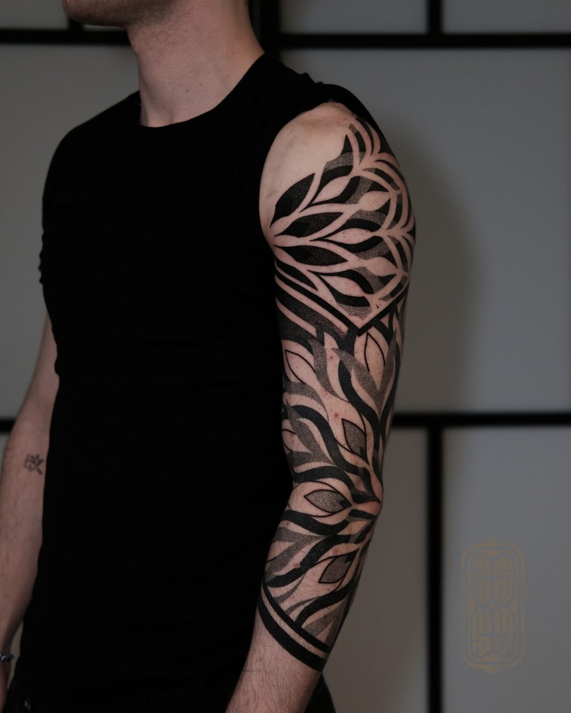 A close-up of a man's arm with a detailed black and grey tattoo featuring intricate leaf patterns.