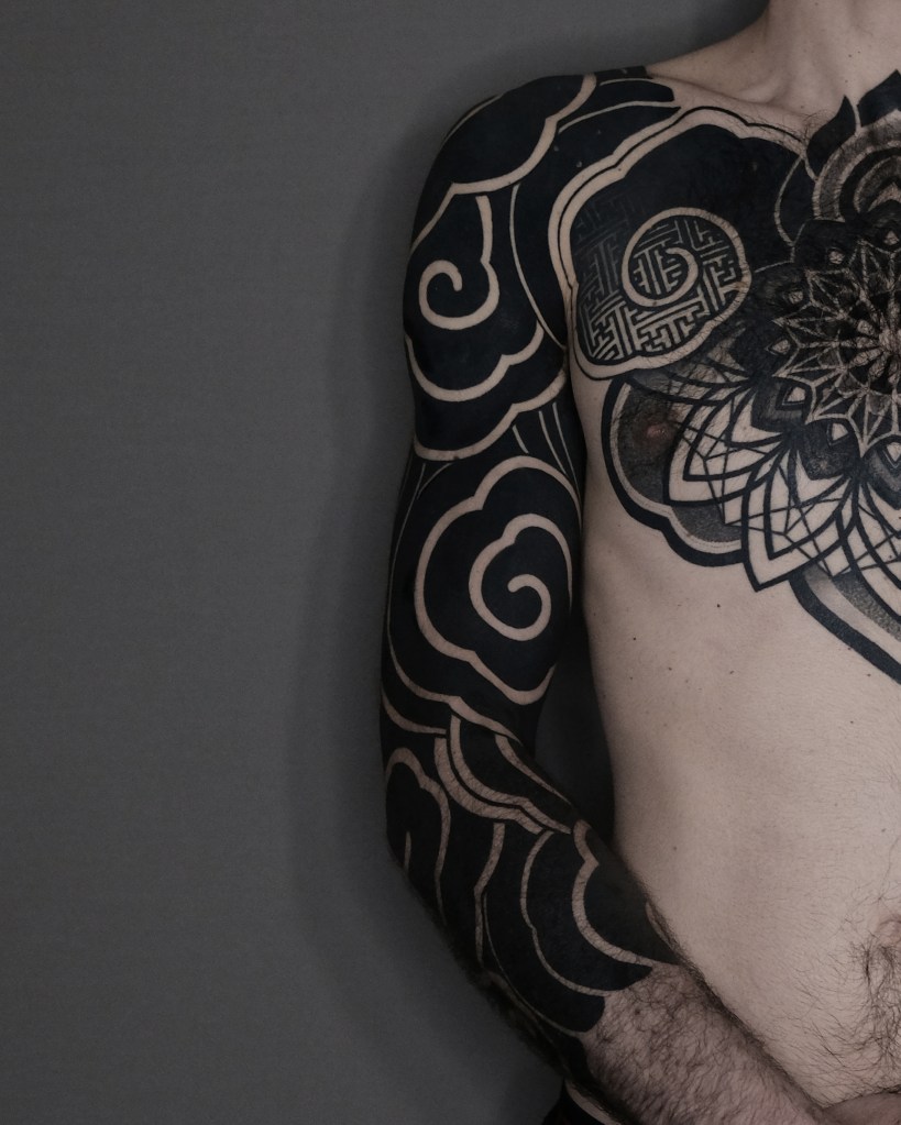 Close-up of a tattooed male torso featuring intricate black and grey designs, including swirling patterns and a mandala.