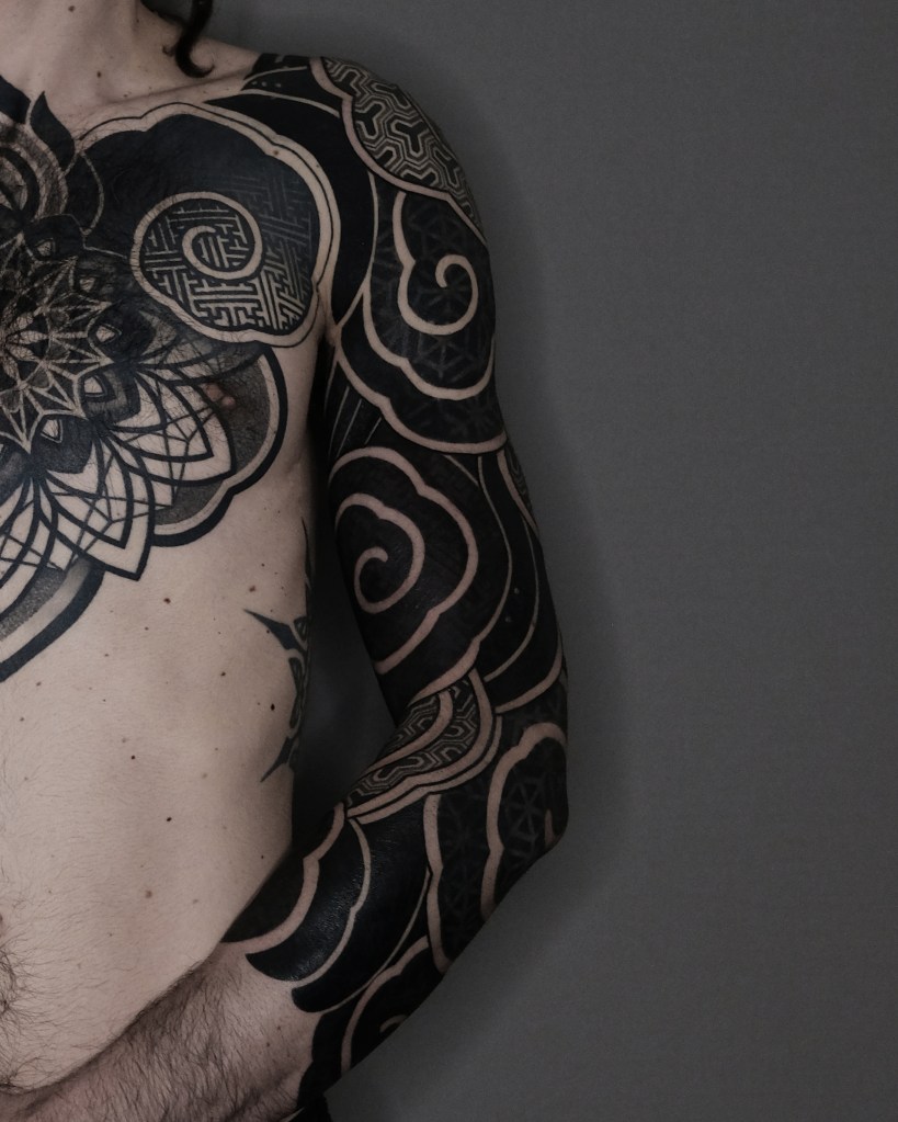 Close-up of a bare torso showcasing intricate black tattoos on the chest and arm, featuring geometric patterns and swirling designs against a gray background.