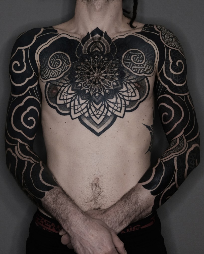 A close-up of a person's torso showcasing intricate black tattoos on the chest and arms, featuring geometric patterns and swirling designs.